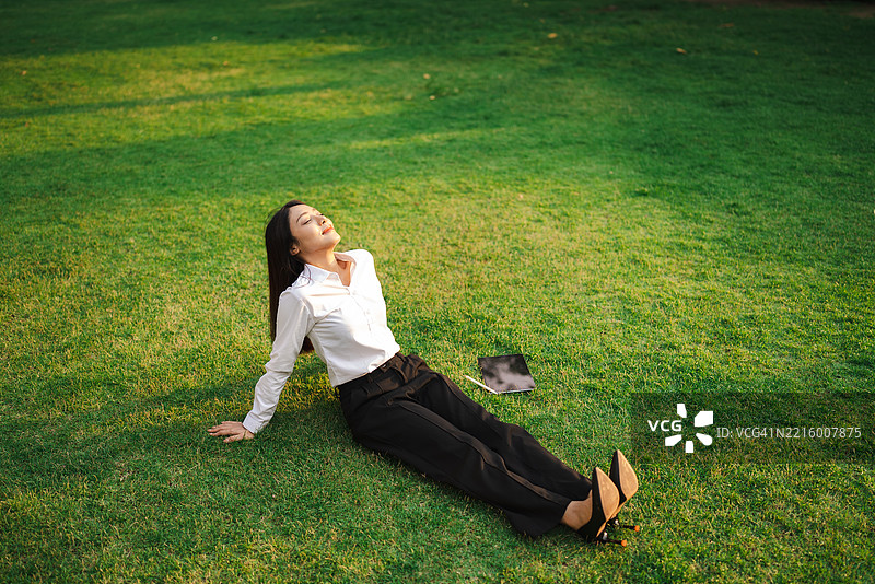 年轻职业女性在郁郁葱葱的草坪上放松，享受阳光，面带宁静的微笑——捕捉忙碌日子间完美的宁静时刻。图片素材