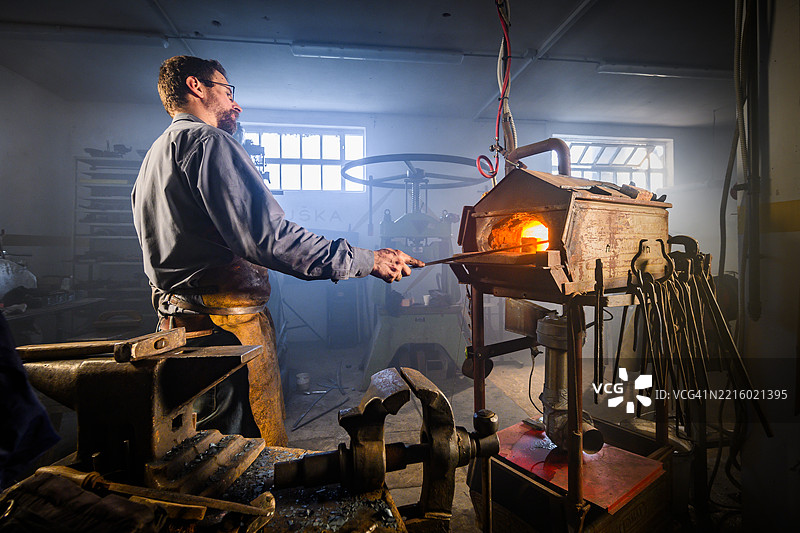 中年男性铁匠在室内工作坊中加热金属，周围有工具和温暖的灯光图片素材