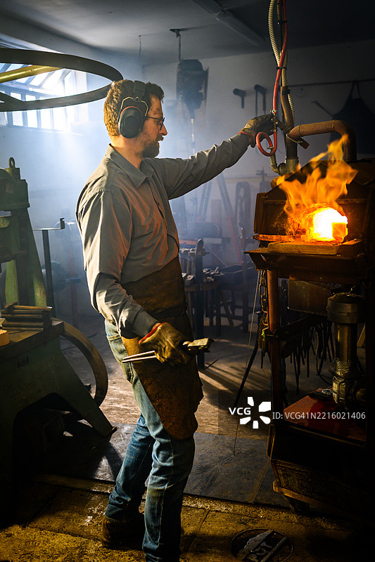 熟练的男性铁匠在昏暗的工作坊中加热金属，火炉发出微光图片素材