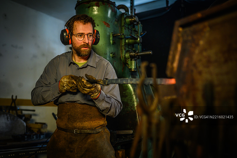 专注的男性铁匠在昏暗的工业熔炉中用钳子加热金属图片素材