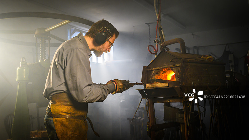 穿着皮围裙的传统铁匠在昏暗的工作坊中于发光的熔炉前工作图片素材