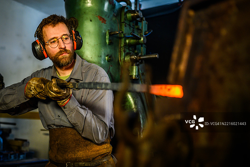 中年黑smith穿着灰色工作服和皮革围裙在工业锻造车间工作图片素材