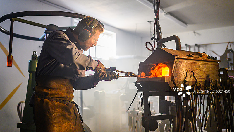 身穿皮围裙的熟练铁匠在传统锻造车间中处理热金属图片素材