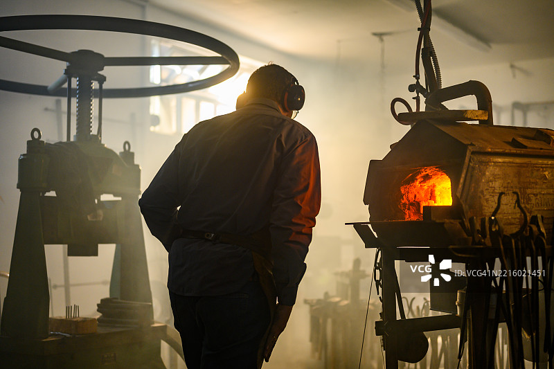 男性铁匠在工业车间的熔炉中加热金属，温暖的光辉照耀着他。图片素材