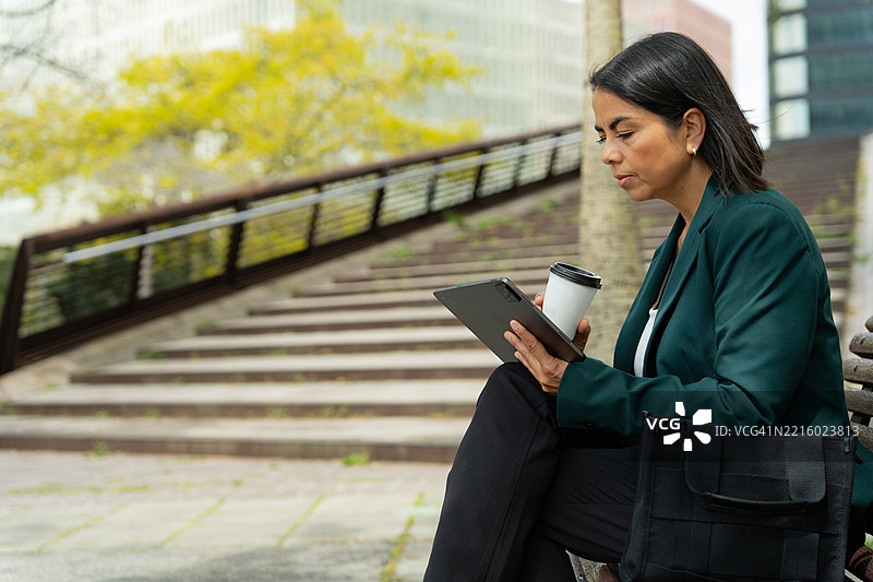 专业女性在城市长椅上远程工作，手持平板和咖啡杯图片素材