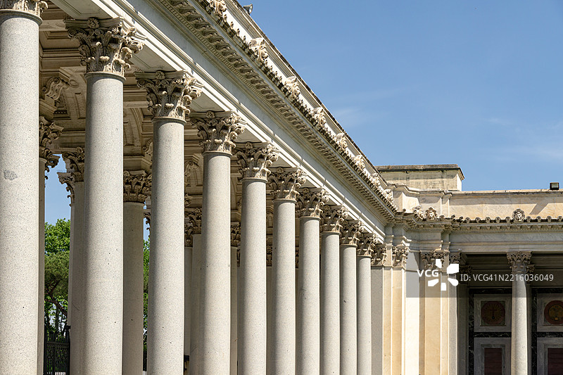 一排宏伟的古典柱子，华丽的柱头支撑着壮丽的檐口，四周环绕着郁郁葱葱的绿植，沐浴在阳光下，映衬着清澈的蓝天，展现出永恒的户外景致。图片素材