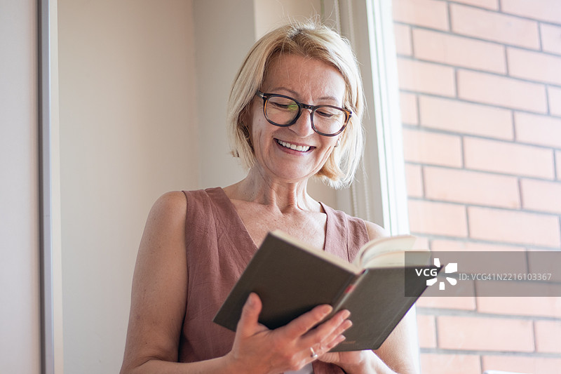一位成熟的金发女性戴着眼镜，站在房间的窗边阅读一本书。图片素材