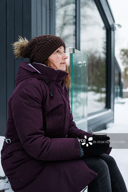 穿着冬季外套的女人在雪天的户外，手里拿着杯子，靠近一栋房子。图片素材