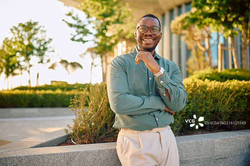 微笑的商人在办公楼旁摆姿势，穿着绿色衬衫和米色裤子图片素材