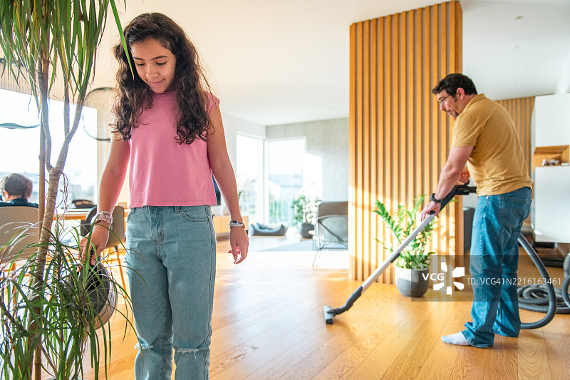 中年西班牙裔父亲正在吸尘，女儿快乐地浇水植物图片素材
