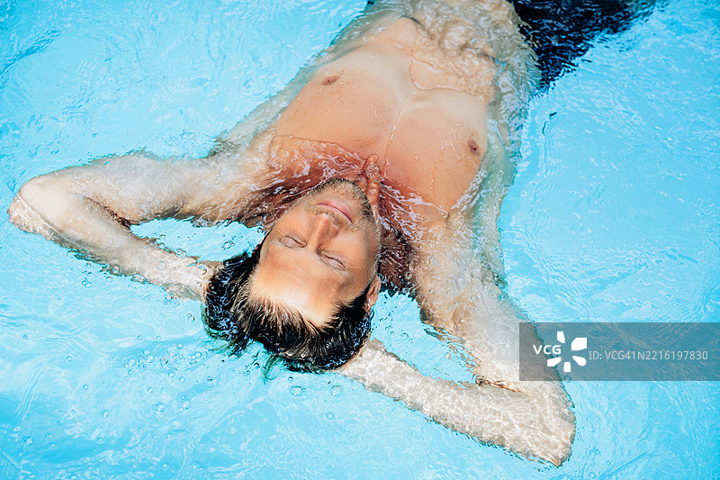 从上方俯视一位英俊的中年男子仰卧在泳池中，双眼闭合，双手放在头后，周围水面微波荡漾。图片素材