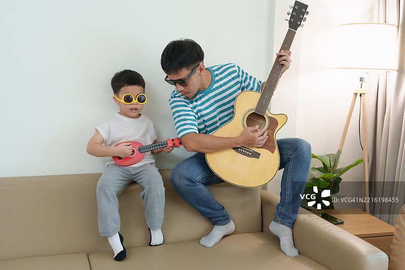 亚洲父子在家中一起享受音乐表演，快乐地弹奏吉他和尤克里里，欢声笑语不断。图片素材