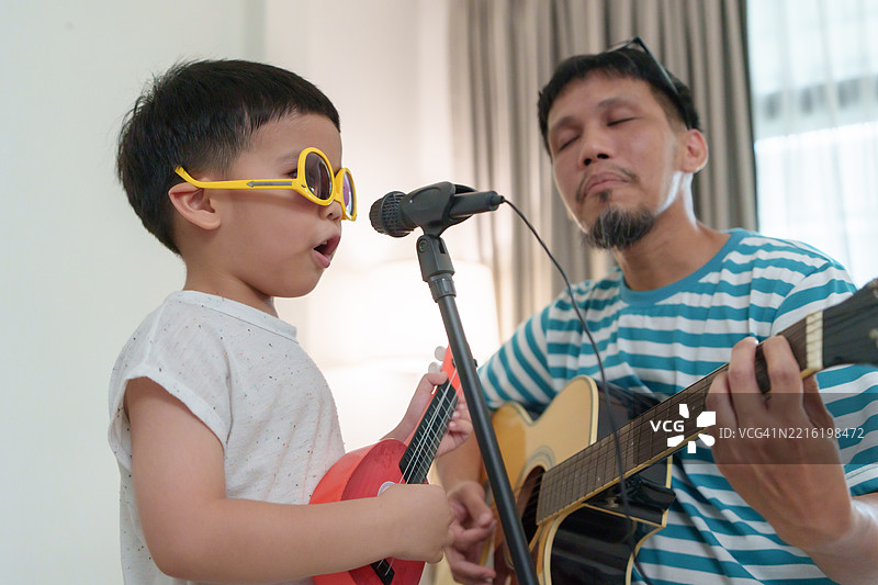 亚洲父子在家中一起表演，男孩快乐地唱歌并弹吉他，享受音乐的美好时刻图片素材