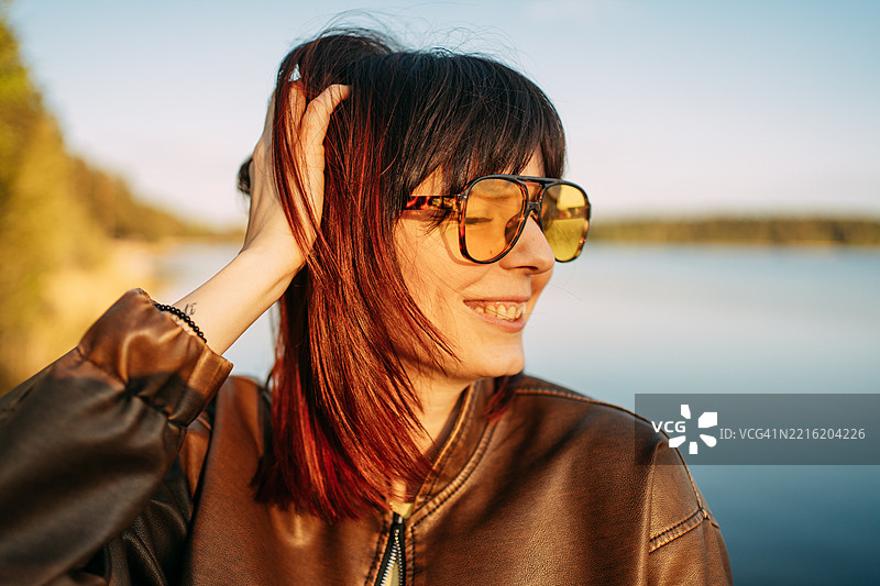 年轻女子在日落时分，戴着黄色太阳镜，轻抚着湖边的头发。图片素材