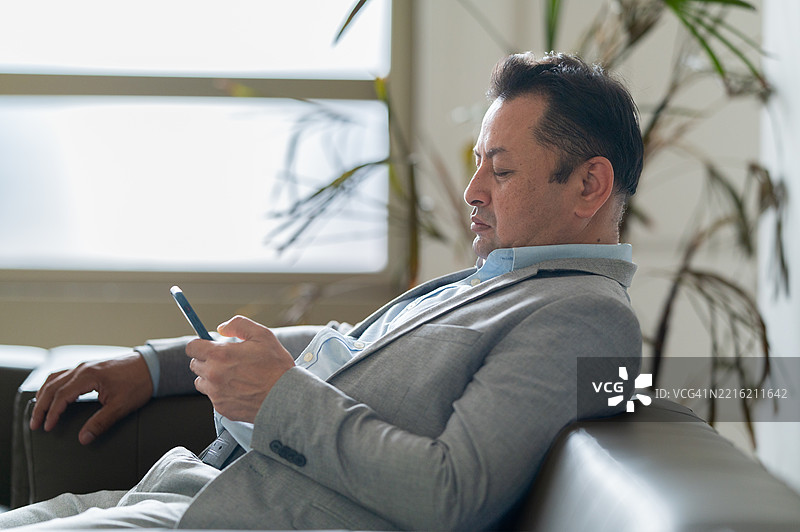 亚洲成熟男性坐在沙发上，手持智能手机，因工作压力过大而睡着的照片图片素材