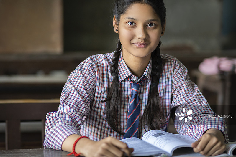 坐在教室桌子上的女学生，手里拿着书，面带微笑看向镜头。图片素材