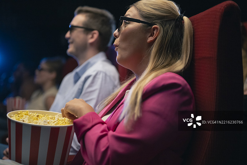 年轻情侣在黑暗的电影院里享受3D电影，手里拿着爆米花，佩戴着特殊眼镜图片素材
