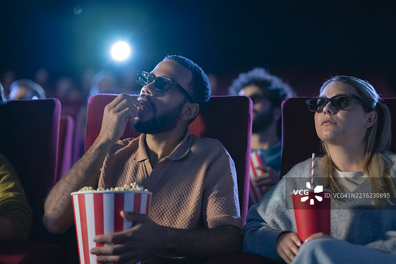年轻多元化情侣在黑暗的电影院观看3D电影，手里拿着爆米花和饮料图片素材