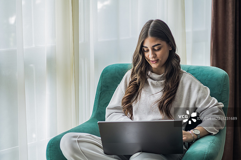 年轻专注的女性在家窗边使用笔记本电脑。在线教育。图片素材