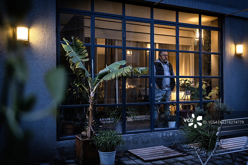 成熟男子在黄昏时分透过现代住宅的窗户向外望去图片素材