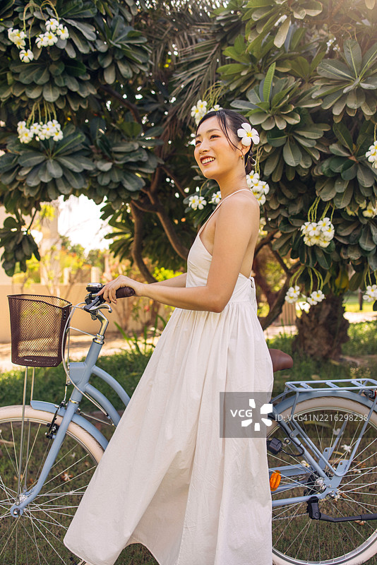 微笑的女性穿着白色连衣裙，骑着自行车在盛开的花树下。图片素材