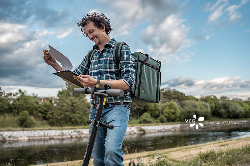 阳光明媚的日子里，背着背包的快递员在风景如画的河边查看文件图片素材