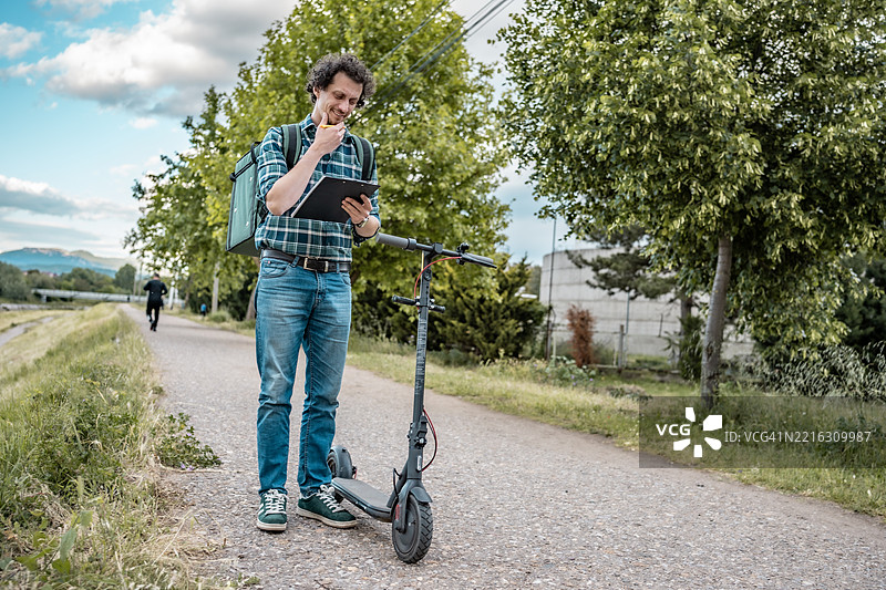 户外阳光明媚的日子里，骑电动滑板车的外卖骑手正在使用平板电脑图片素材
