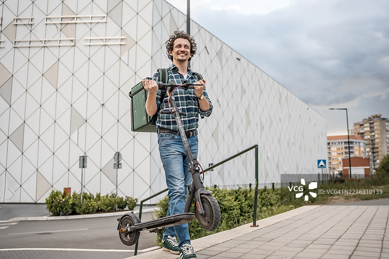 男子在现代城市建筑外微笑着骑着电动滑板车图片素材