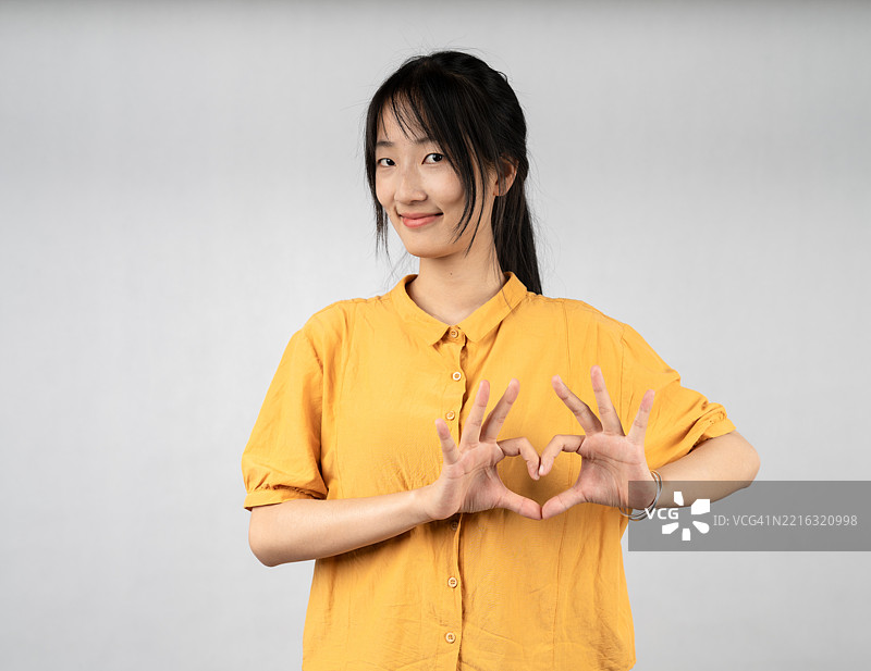 穿着黄色衬衫的年轻女性用手做出心形手势，营造出愉快友好的氛围图片素材