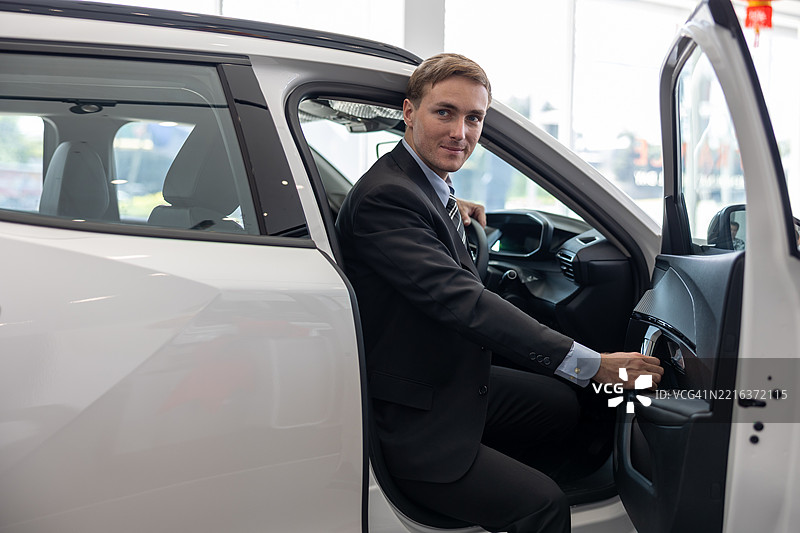 一位自信的商人在一家汽车经销商的新白色SUV驾驶座上坐着，转身面对镜头，同时评估车辆的内部功能。图片素材