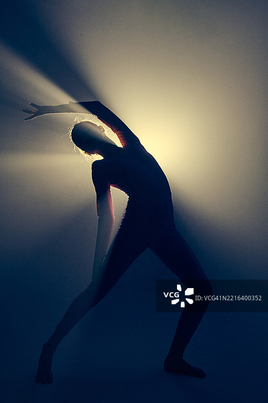 一位身穿紧身衣的体操女孩在黑暗背景下的剪影中进行体操动作。光线射线。图片素材
