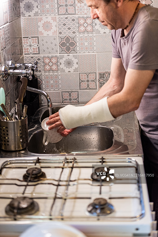 受伤的男子左手骨折，包裹在石膏中，在家洗碗图片素材