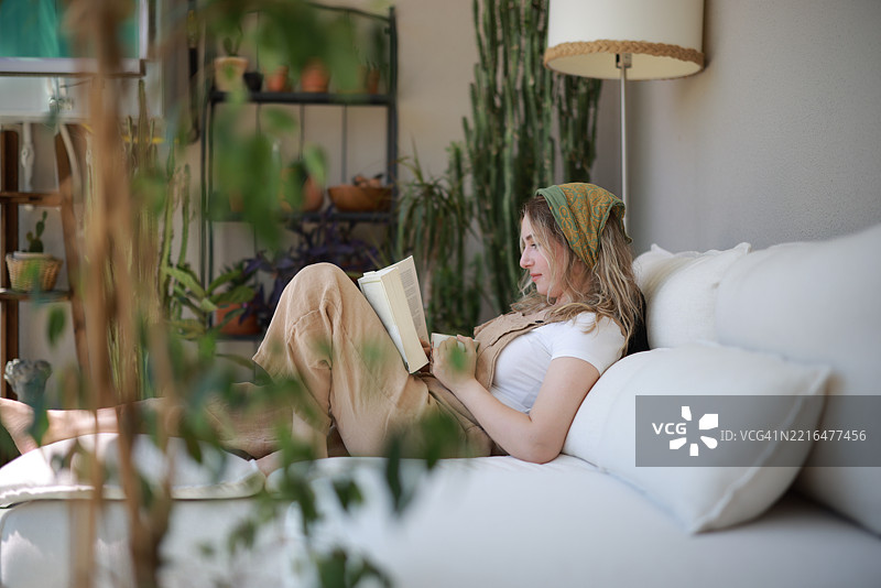 年轻女性穿着工装，坐在沙发上阅读一本书，喝着咖啡图片素材