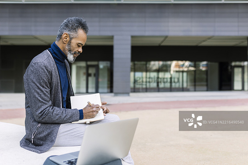 商人在建筑物前坐着，边写日记边使用笔记本电脑。图片素材