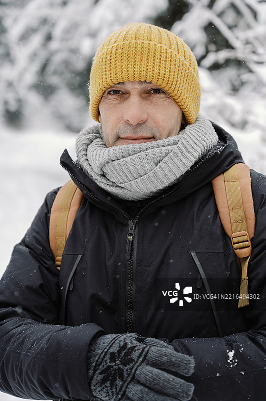 成熟男性戴着黄色帽子，灰色围巾，黑色夹克，背着橙色背包，站在雪-covered森林中。图片素材
