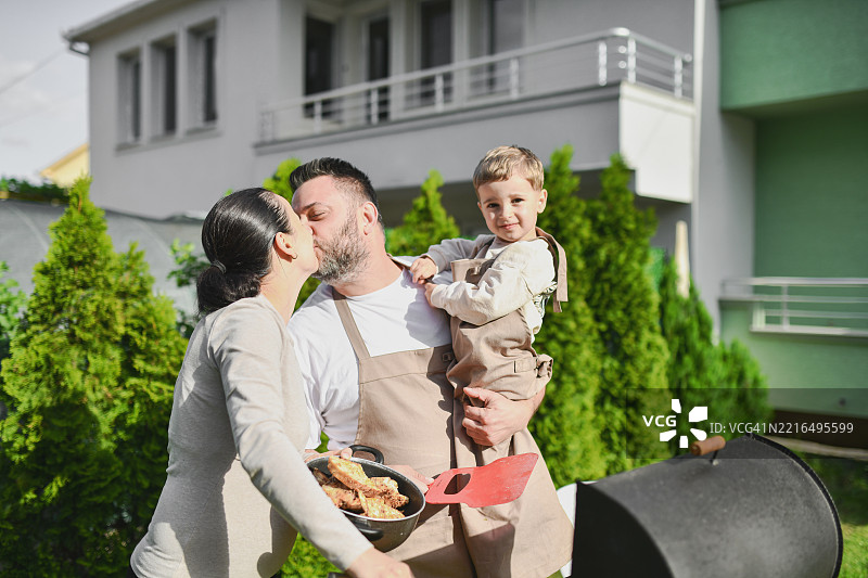 温馨的家庭时刻，父母亲吻，父亲抱着幼儿，正在准备花园烧烤。图片素材