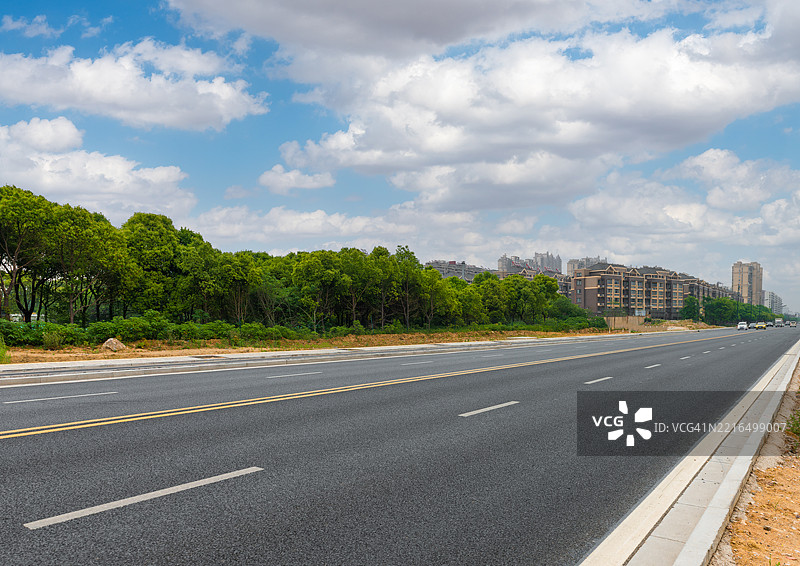 天空晴朗，白云飘浮，沥青道路延伸向远方，路两旁是绿色的树林。图片素材