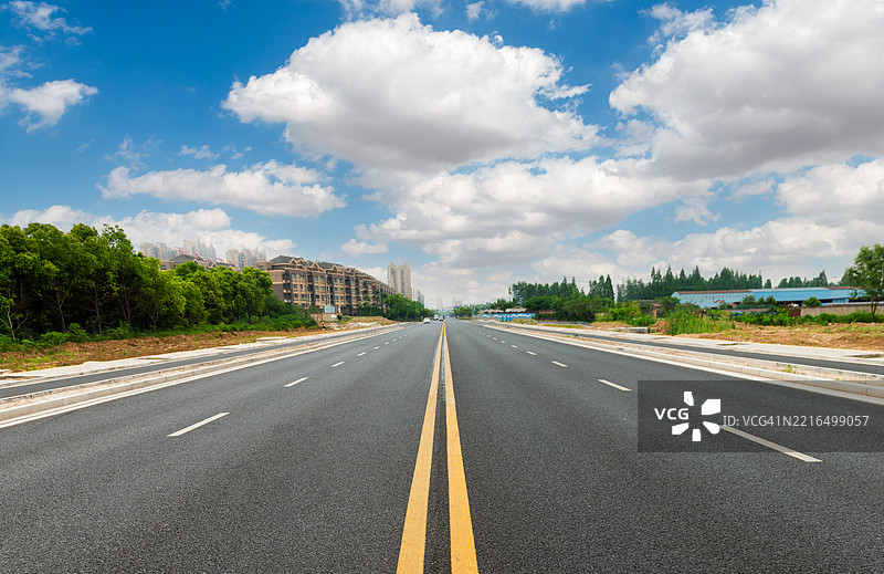 天空晴朗，白云飘浮，一条沥青路通向远方，路两旁是绿色的树林。图片素材