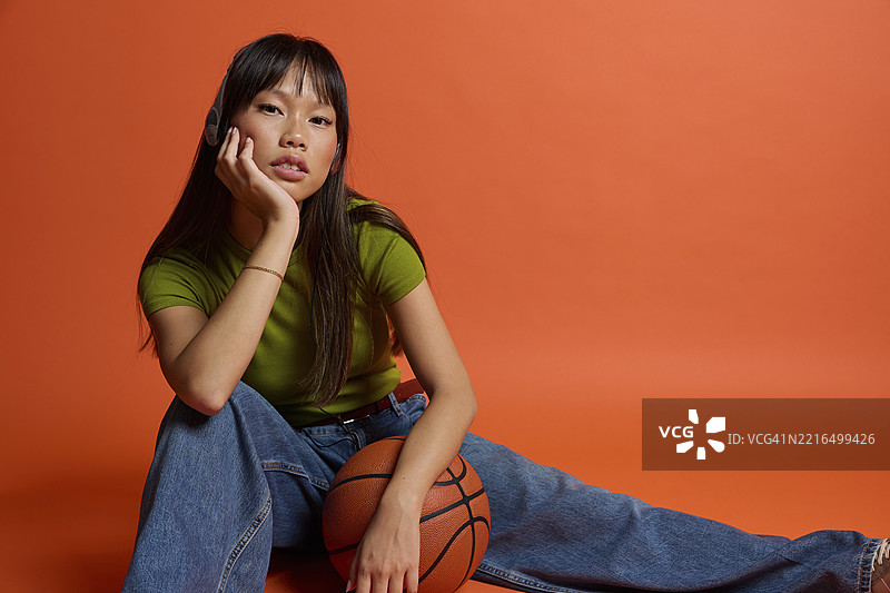 年轻女性坐在橙色背景前，手里拿着篮球图片素材