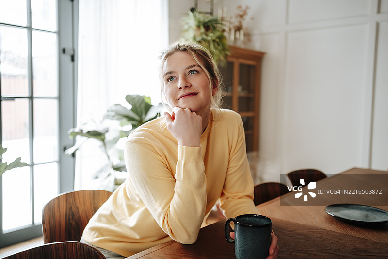 穿着黄色毛衣的女人坐在桌子旁，手里拿着一个杯子，神情沉思。图片素材