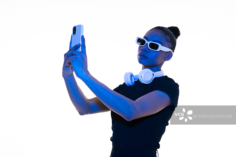 在工作室里，佩戴太阳镜和无线耳机的人在蓝光下使用智能手机图片素材