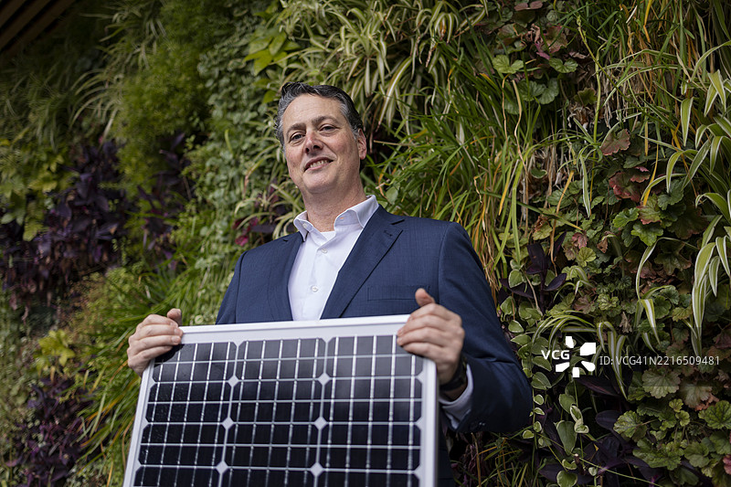 男子在户外绿色墙前手持太阳能电池板图片素材