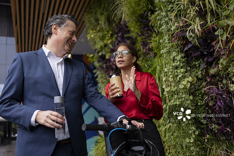 商务同事在户外绿墙旁谈话，旁边有一辆滑板车和咖啡。图片素材