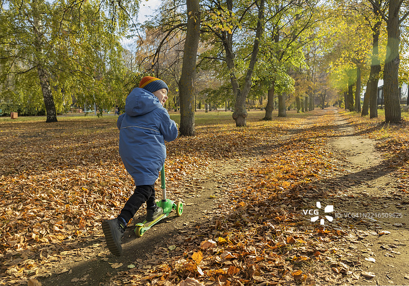 男孩在秋天的公园里骑滑板车，周围是落叶和阳光。图片素材