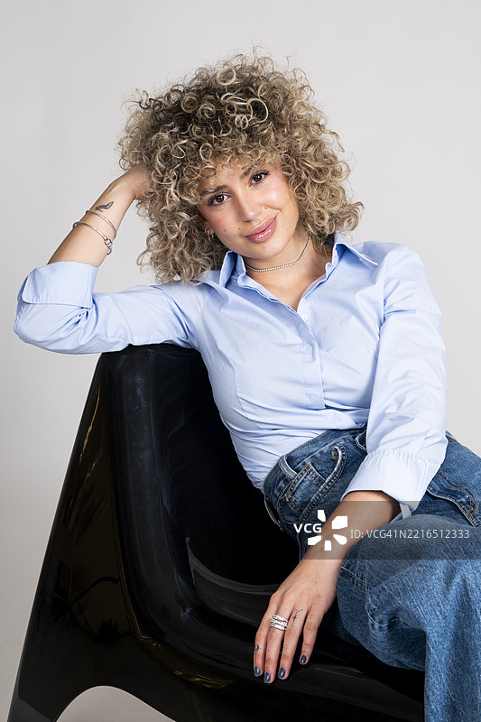 时尚的卷发女性坐在工作室的椅子上，穿着休闲服，自信地看着镜头。图片素材
