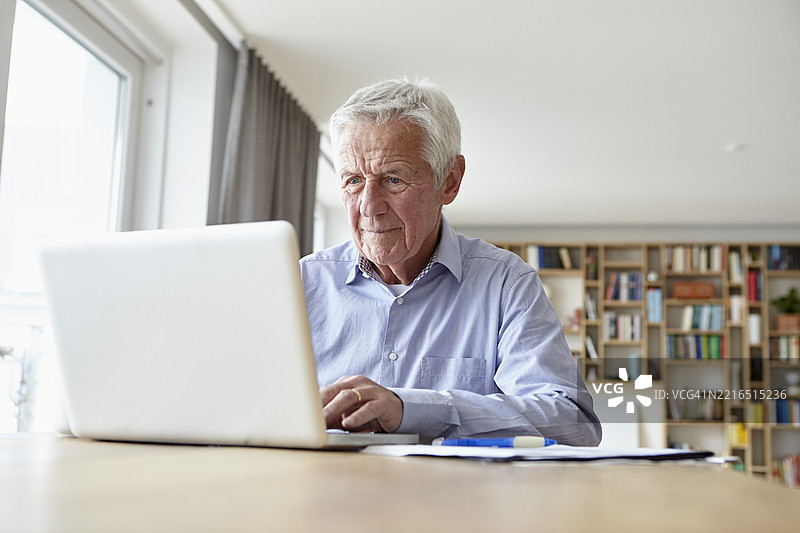 一位老年男子在家中使用笔记本电脑，背景有文件和书架。图片素材