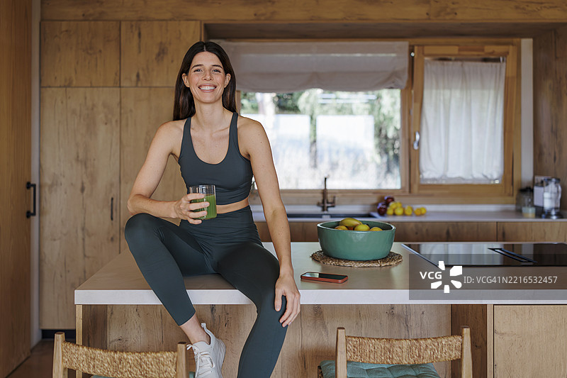穿着运动服的女性坐在厨房台面上，手里拿着绿色果汁。图片素材