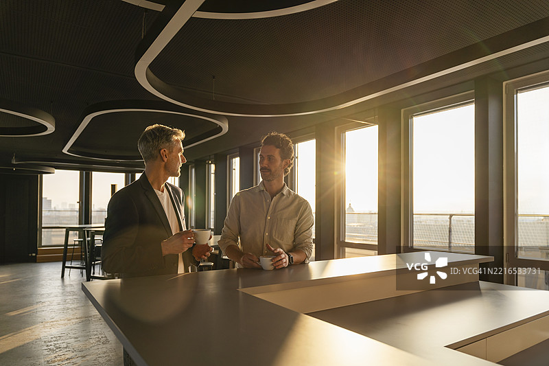 两名男子在现代办公室的日落时分举行商务会议，手中拿着咖啡。图片素材