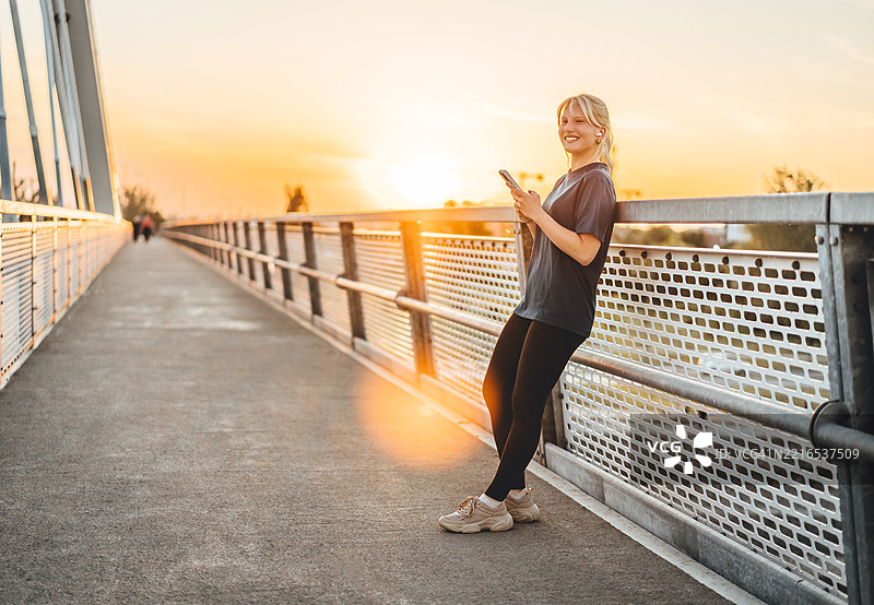 年轻的运动女性在日落时分的桥上使用智能手机图片素材
