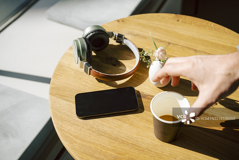 手伸向咖啡杯，桌上有耳机和智能手机，咖啡馆内景图片素材